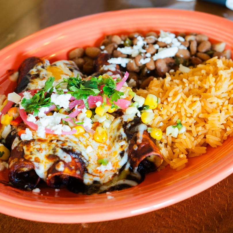 Enchiladas topped with cheese, cilantro, and pickled onions, served with rice and beans on an orange plate.
